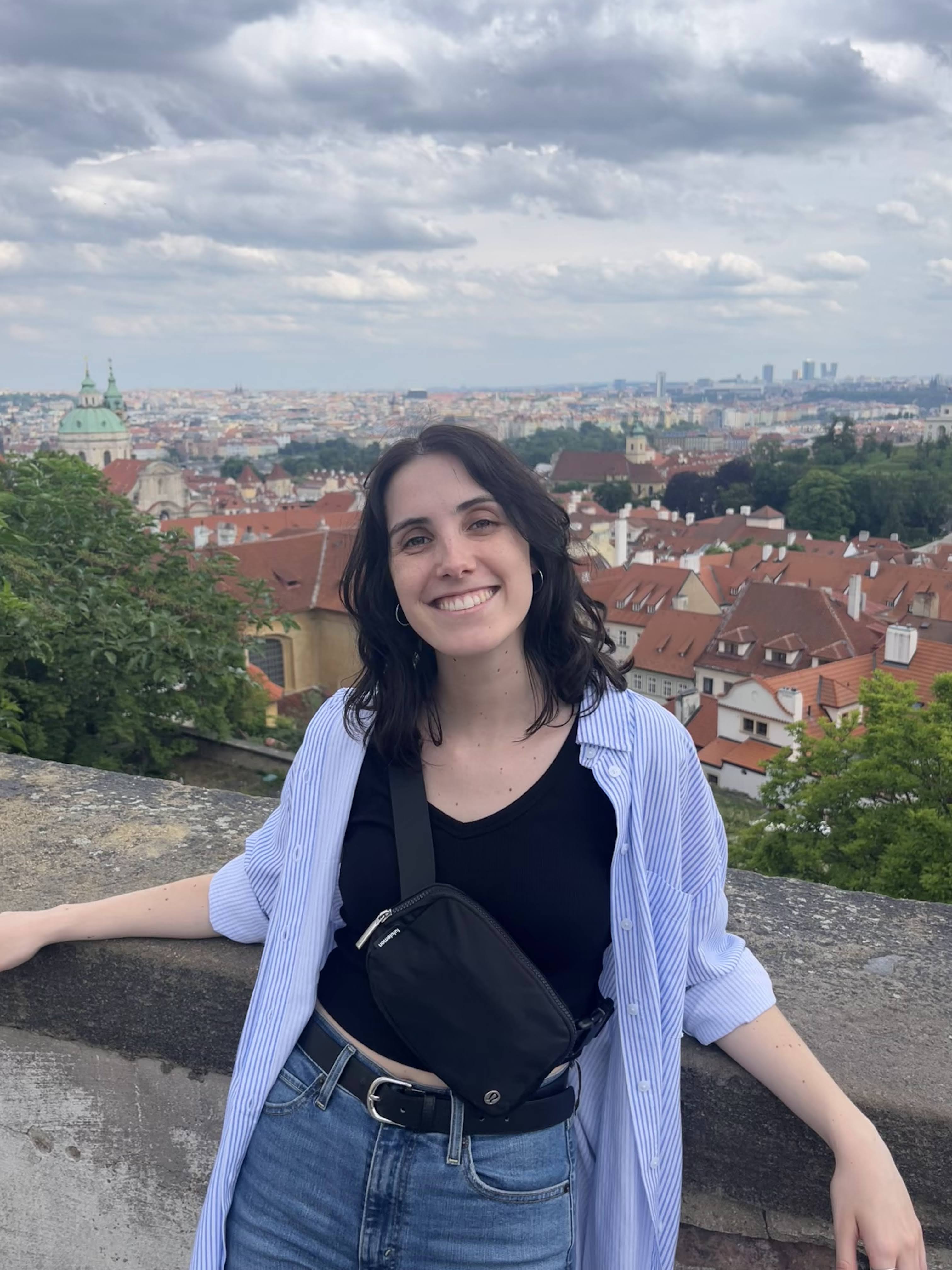 Mireia has shoulder-long dark brown hair and dark brown eyes, and light skin. She wears blue jeans with a black belt, a black tailored shirt, and a loose open shirt with thin white and blue stripes over it. A small black bag hangs diagonally over her upper body. Mireia smiles widely into the camera. She leans casually against an old wall with her arms resting on the top of the wall. In the background you can overlook a European-looking city showing houses with red roofs and green-leaved trees, under a clouded sky with the sun lightening up the clouds.