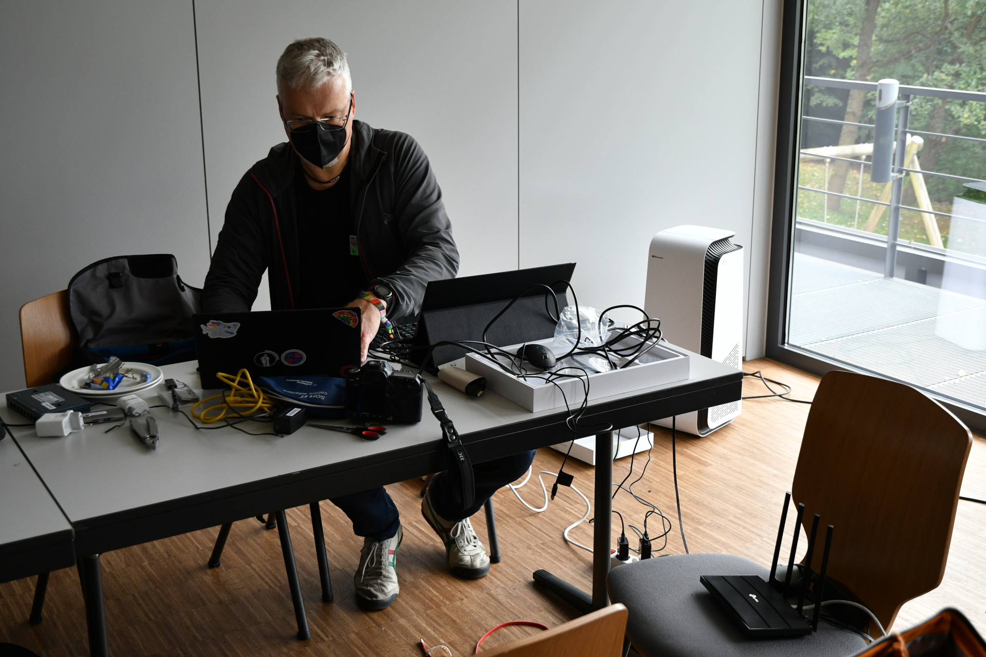 Claudius looking hacky A masked man in dark cloths sits at a table. On the table is a lot of technical equipment like a camera, a laptop, a tablet, some tools, multiple cables for ethernet and power and also a multitool.