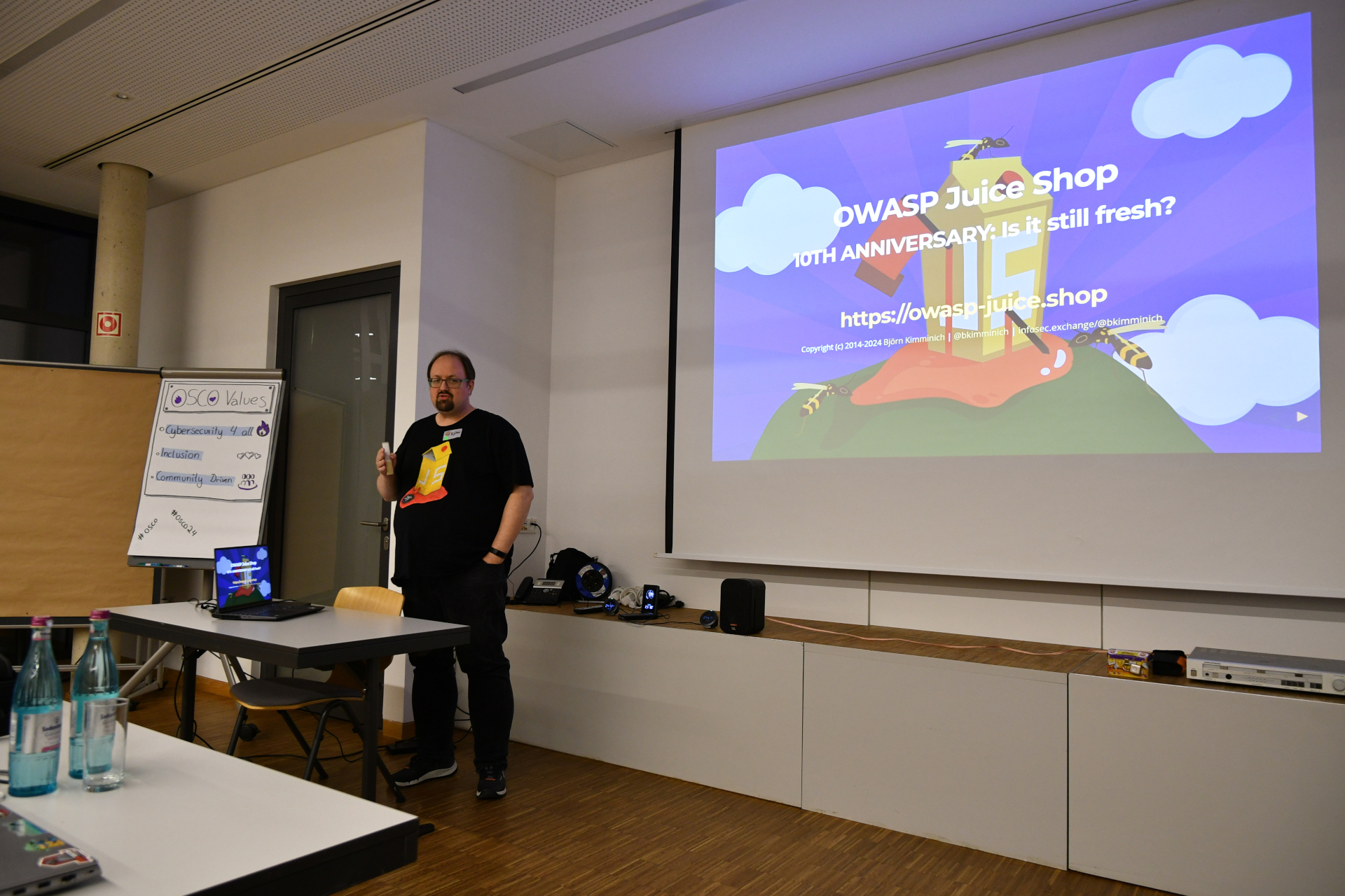 Keynote: OWASP Juice Shop’s 10th Anniversary One of the two keynotes: OWASP Juice Shop’s 10th Anniversary by Björn. Björn stands in front speaking to the audience behind the photograph. On the right site is the first page of his presentation on the wall. On his left site is the osco values flipchart.