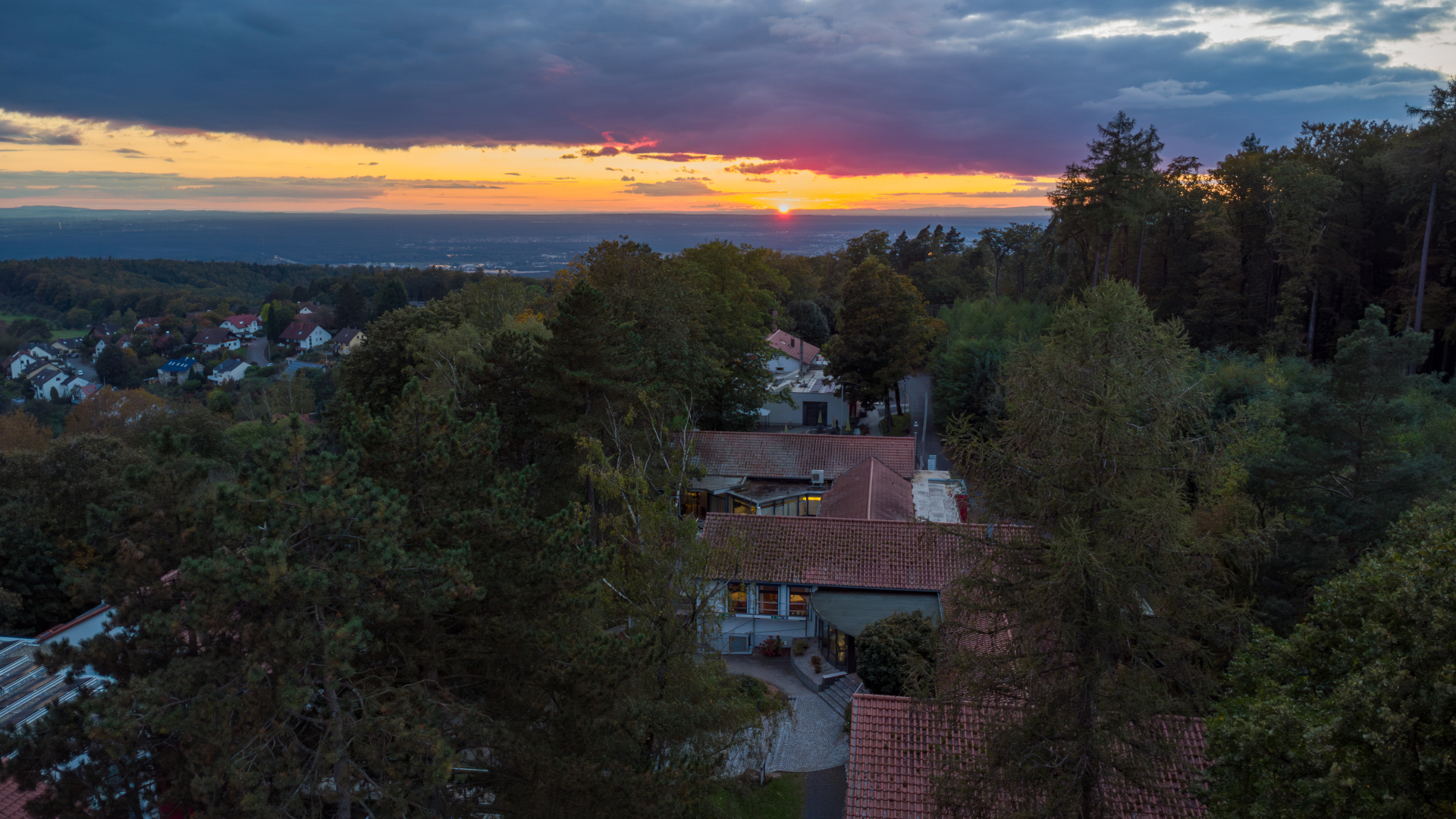 Seminarzentrum at Dusk A drone shot of the Seminarzentrum Rückersbach at dusk. The view is from above the building directed at the settling sun at the horizon. The sky is cloudy and they appear very dark above the sun. The Seminarzentrum is surrounded by trees. On the left and in the background are more houses.