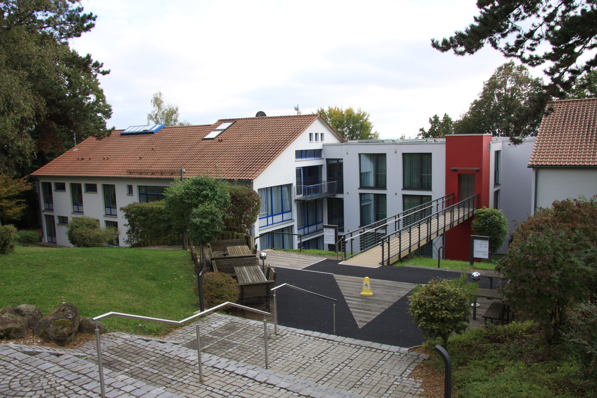 Sleeping Rooms This picture shows two of the buildings with the rooms for visitors of the Seminarzentrum RÜckersbach. A smaller on connects both with an elevator and a bridge to the front there some stairs lead to the photograph. The buildings are surrounded by grass and trees. The sky is cloudy.