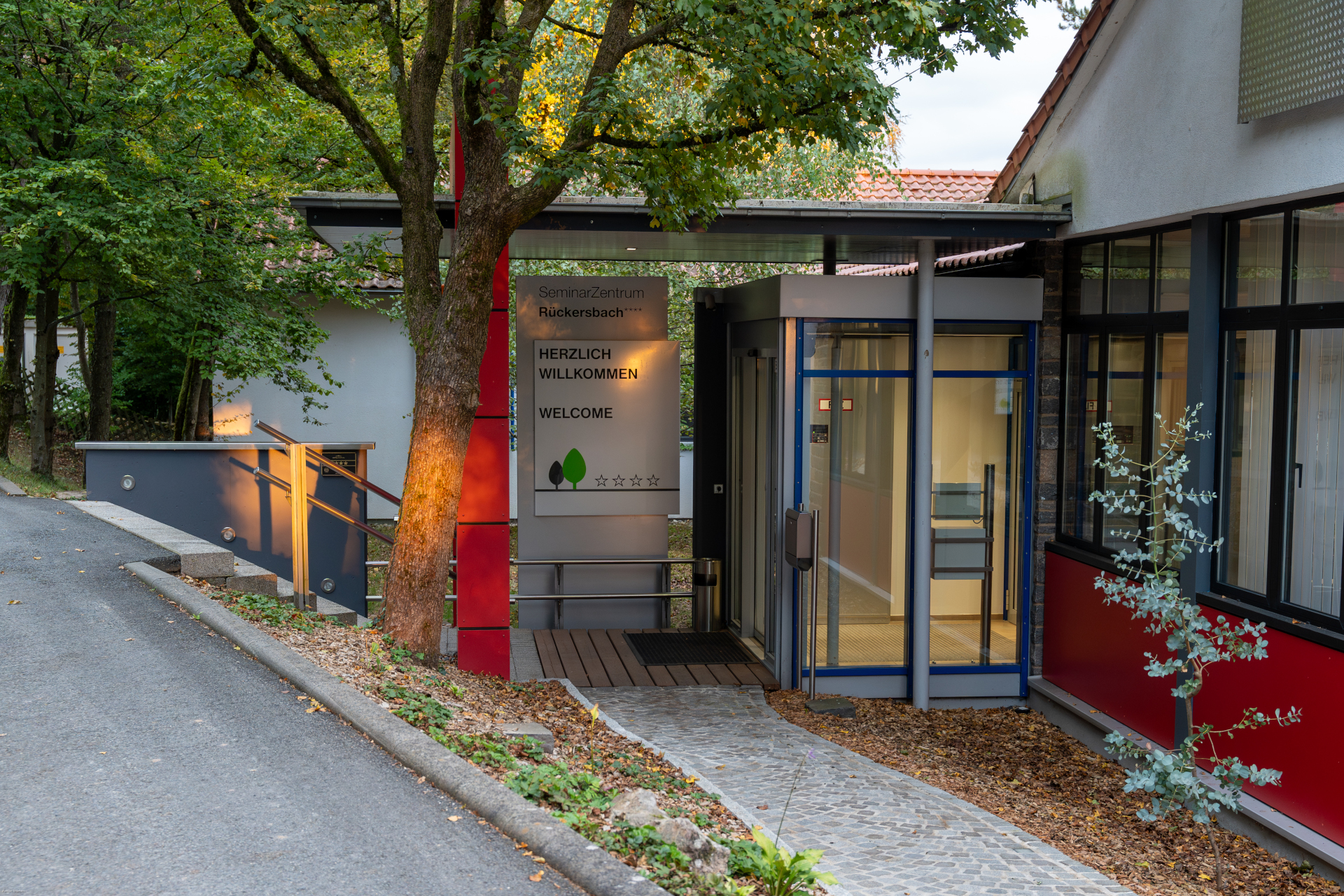 Entrance to the Conference The picture shows the main entrance to the Seminarzentrum Rückersbach, the event location of the osco. Warm sunlight shines through some trees behind the photograph. A sign welcomes the visitors.