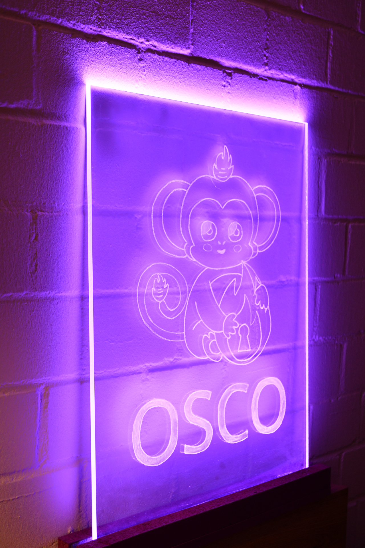 osco (they/them) A photo of the osco logo engraved on a big acrylic glas plate shining in bright purple. The rest of the room is dark and the acrylic glas is standin in front of a brick wall generating nice effects with light and shadow.