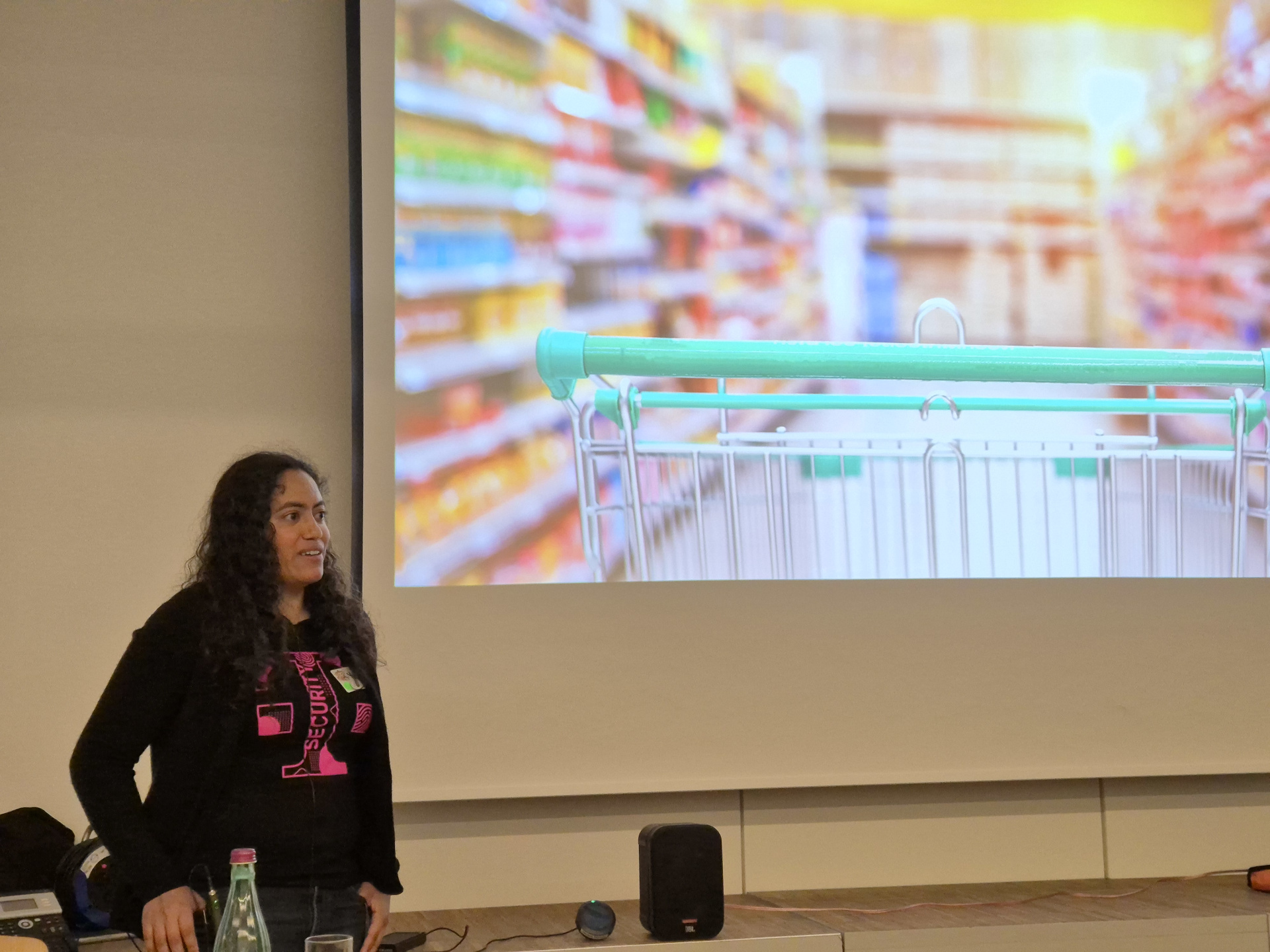 Keynote: How to Hack a Company in a Day One of the two keynotes: Red Teaming by Yvonne. Yvonne stands in the left part of the photo. The most part is a slide of her keynote. A picture of a shopping trolley in a shop.
