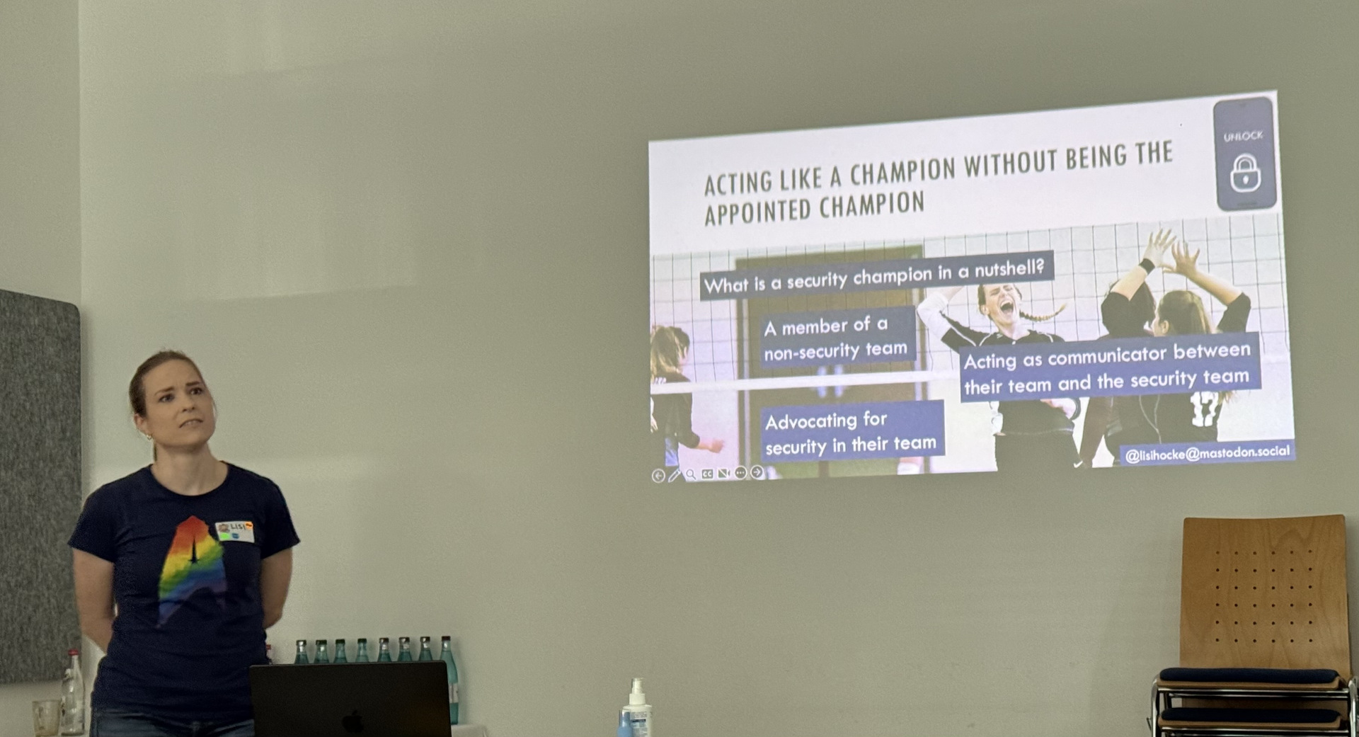 Session Security Champion A woman with black tshirt and rainbow colored Star Trek logo stands with her hands behind her back looking to the audience behind the photograph. To her right is her presentation at the wall. The current slide is titled 'Acting like a champion without being the appointed champion'. The slide is about security champions and their definition.