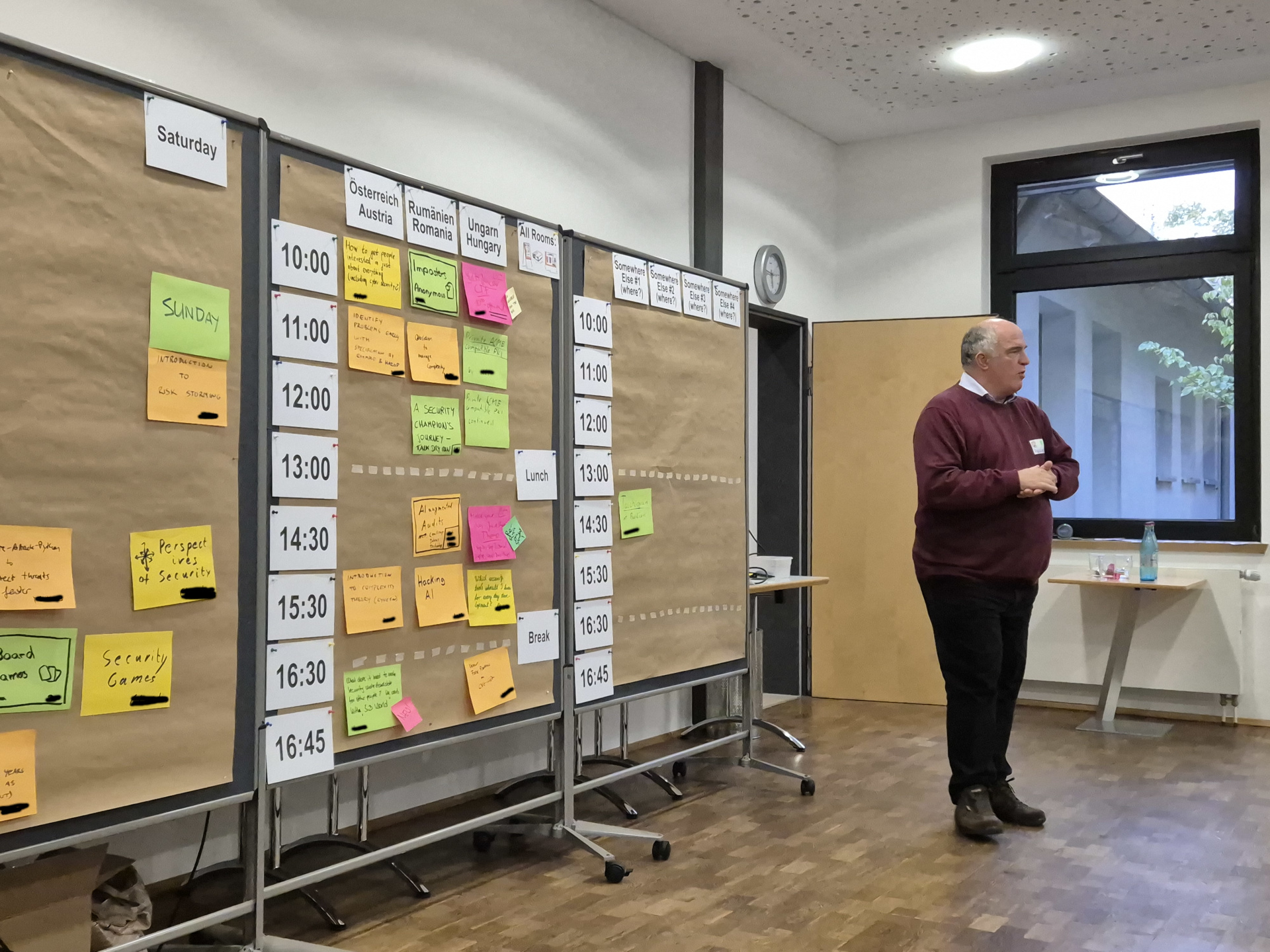 Session Planning A photo the session planning for the first day of osco 2024. On the left wall are parked slots, that could be happening. On the middle wall are the offered sessions in three parallel rooms. On the right wall is the planning for the next day. One sessions is already there. On the right site of the corner stands a man with red sweater explaining. Your can't see the persons he is talking to.