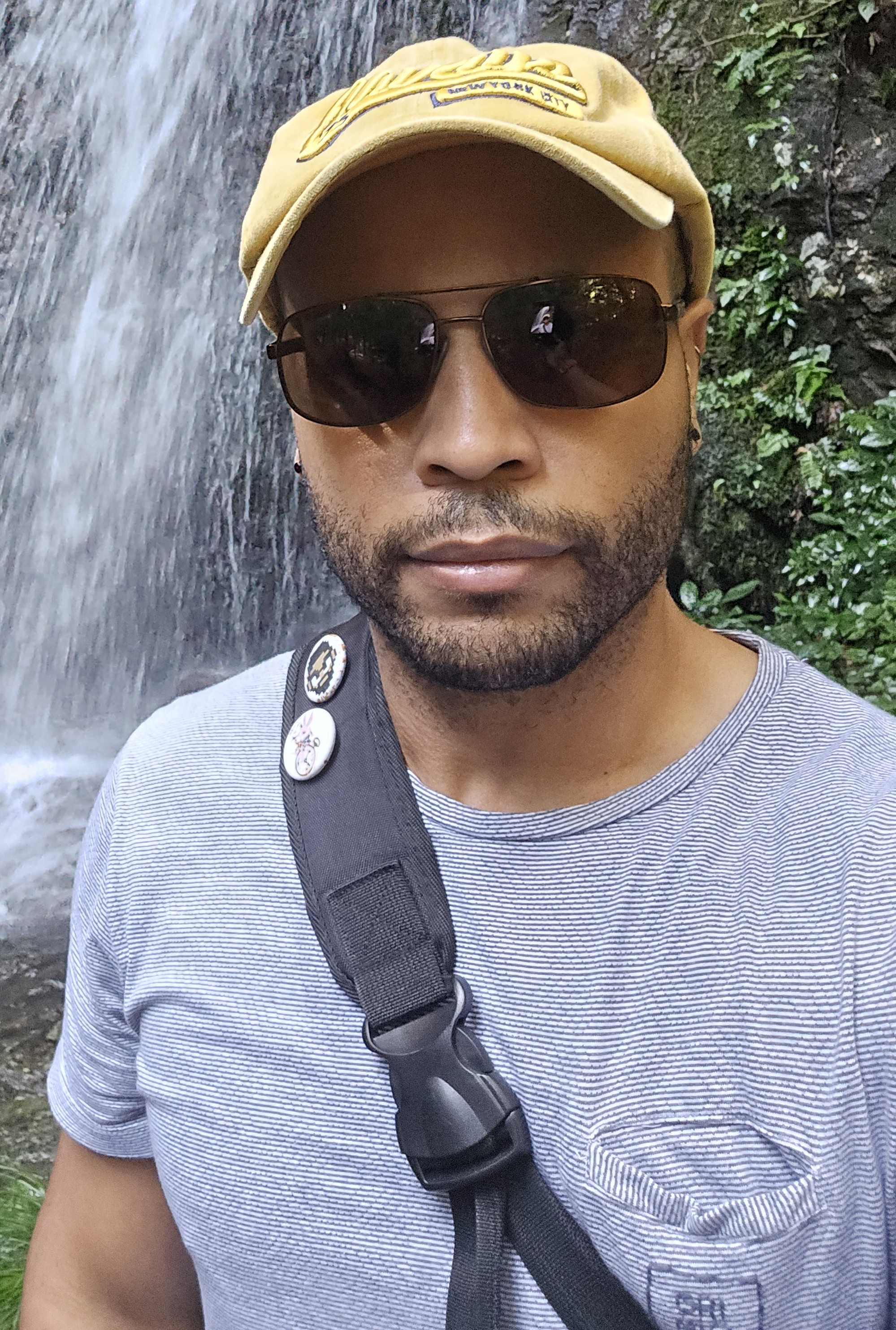 Jahmel has short dark brown hair and wears a short full beard. His dark brown eyes are behind brown sunglasses. He is wearing a light beige flat cap, a T-shirt with thin blue and white stripes, and a messenger bag with two round pins on it. He stands in front of a waterfall with lush green plants on its side.
